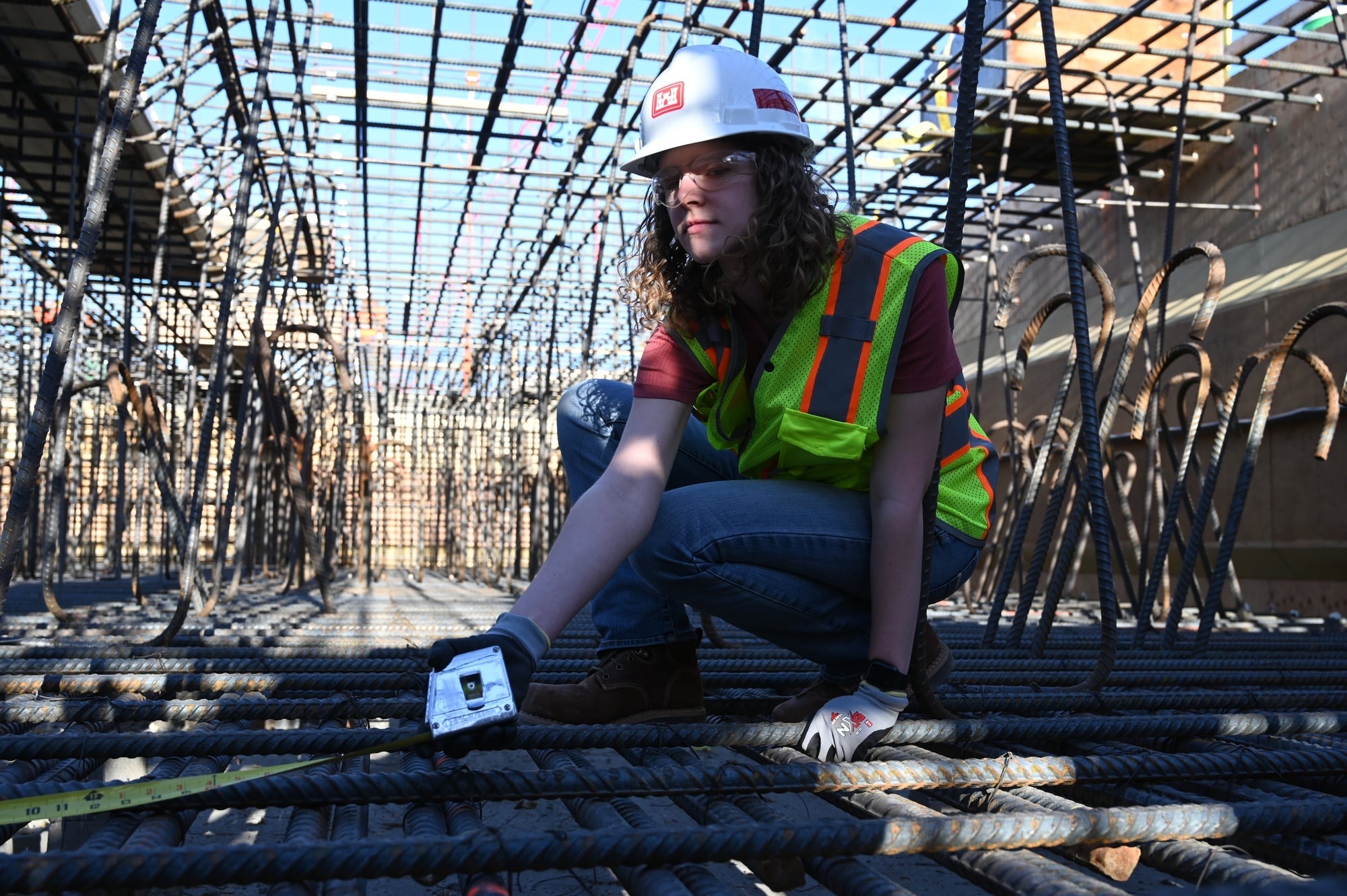A construction worker in a hard hat, safety glasses and hi-vis vest kneels with a tape measure on a dense two-layer rebar mat, with a tunnel of vertical bars and hooked dowels receding behind