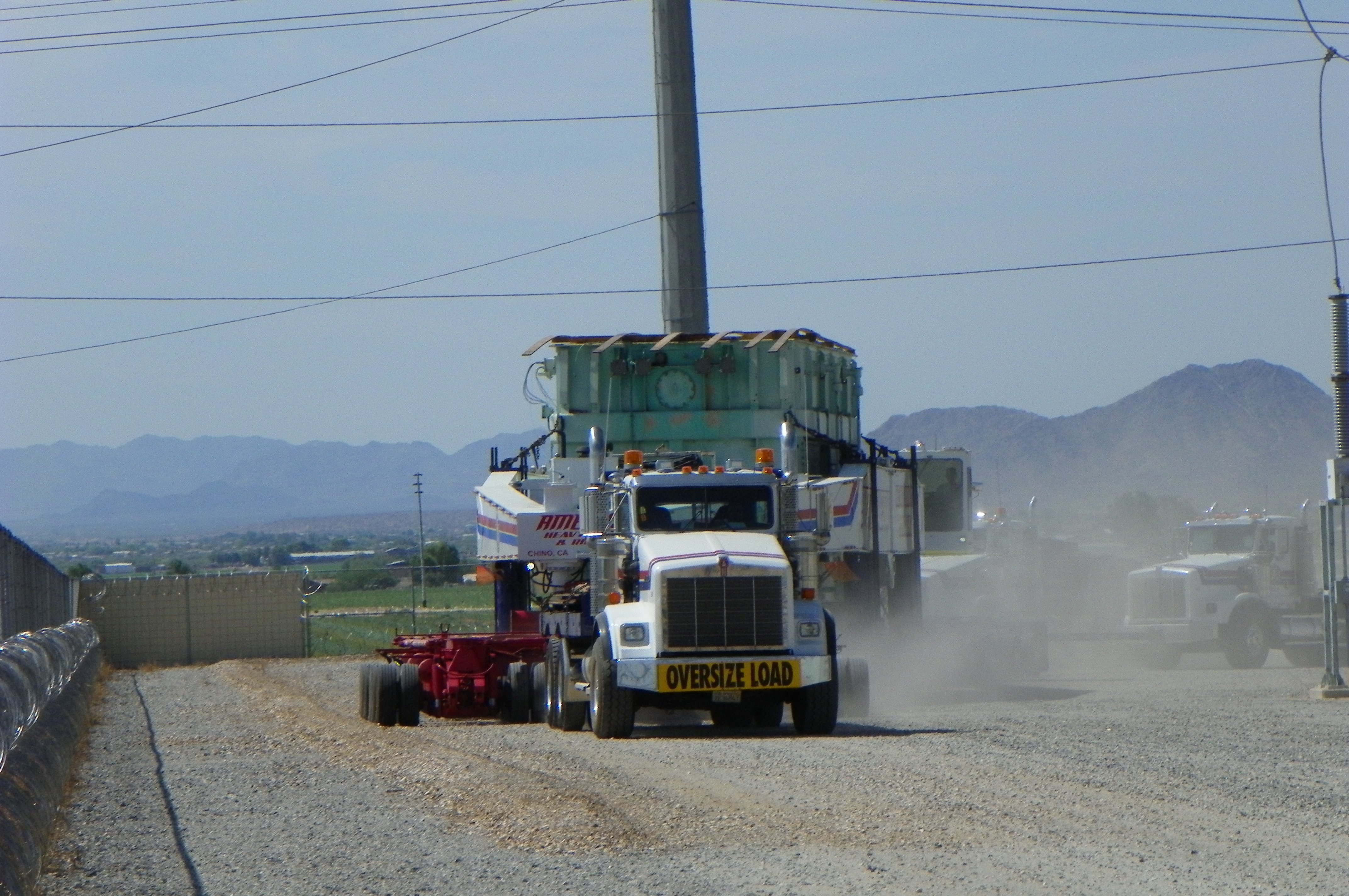 A white heavy-haul truck marked OVERSIZE LOAD hauls a massive green power transformer on a multi-axle trailer down a gravel road, kicking up dust, with desert mountains and escort trucks behind