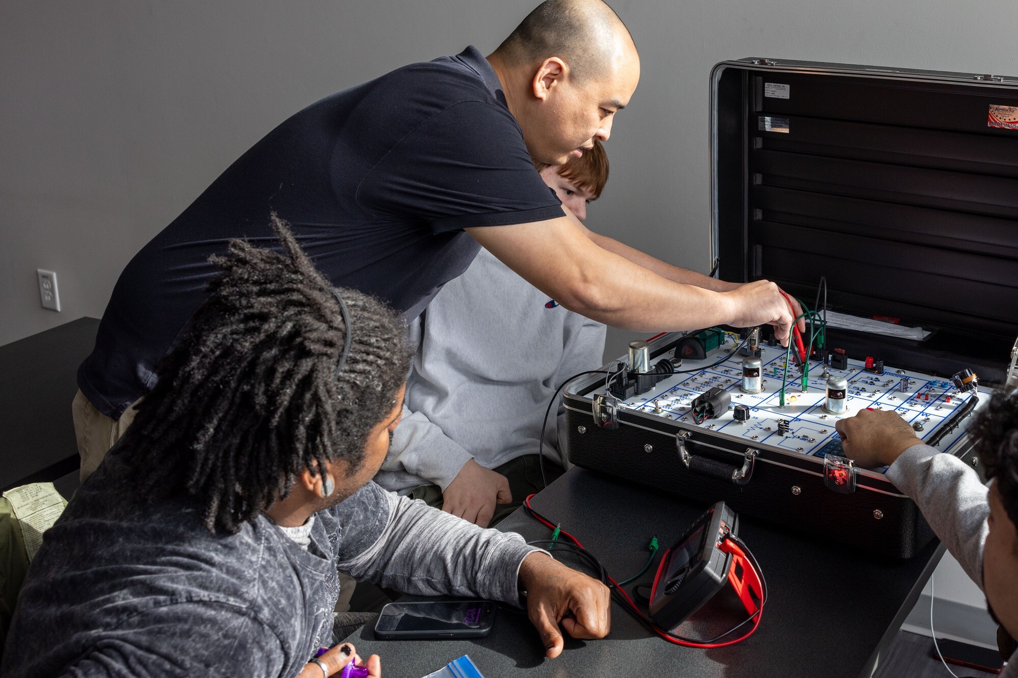 An instructor leans over a wired circuit trainer with a red meter probe while a student beside him watches the board, a multimeter and test leads on the table in front