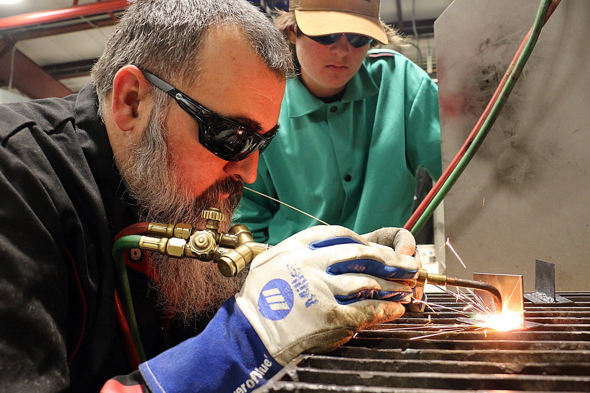 A bearded welding instructor in dark safety glasses guides an oxy-fuel torch across steel as sparks fly, while a student in a green welding jacket leans in over his shoulder to watch