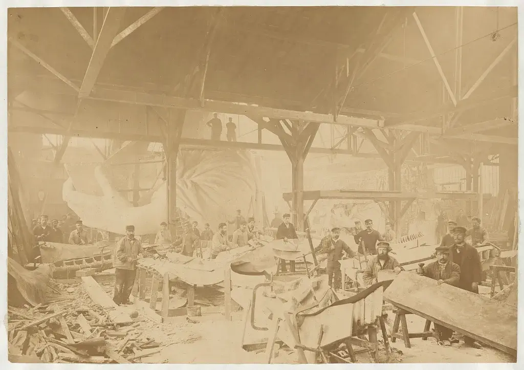 Coppersmiths hammer the Statue of Liberty's copper skin in the Paris workshop, the statue's hand and head behind them.
